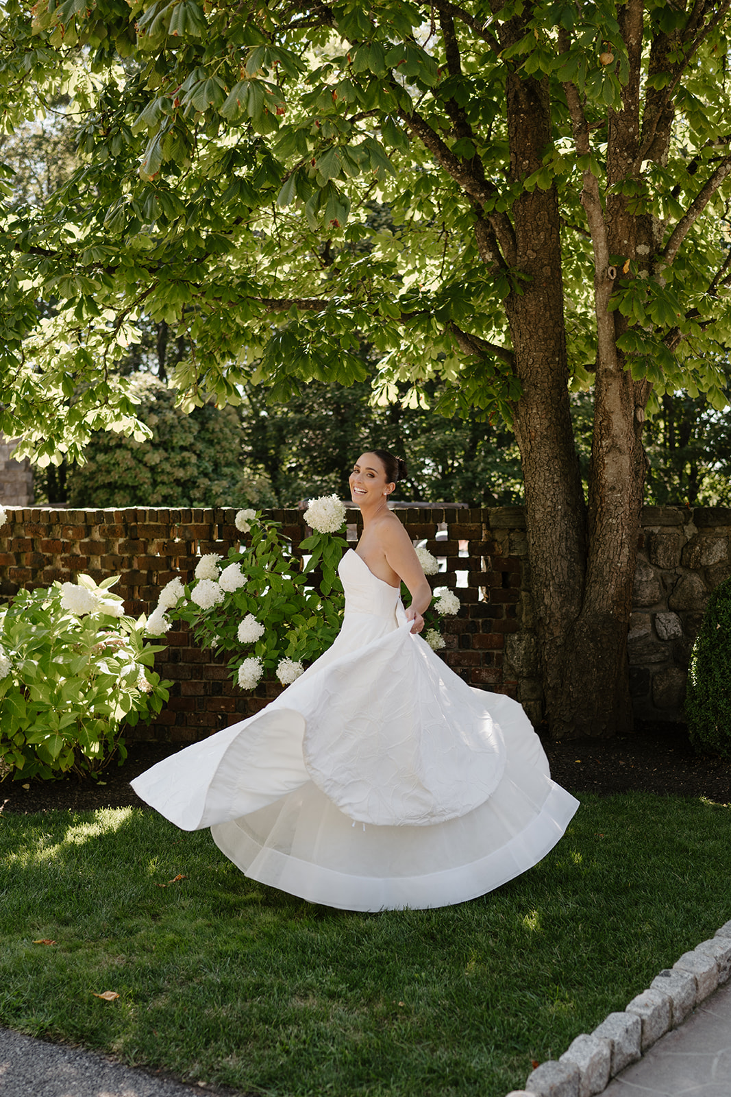 Bride smiling playfully while twirling her gown on the lawn in front of a brick wall.